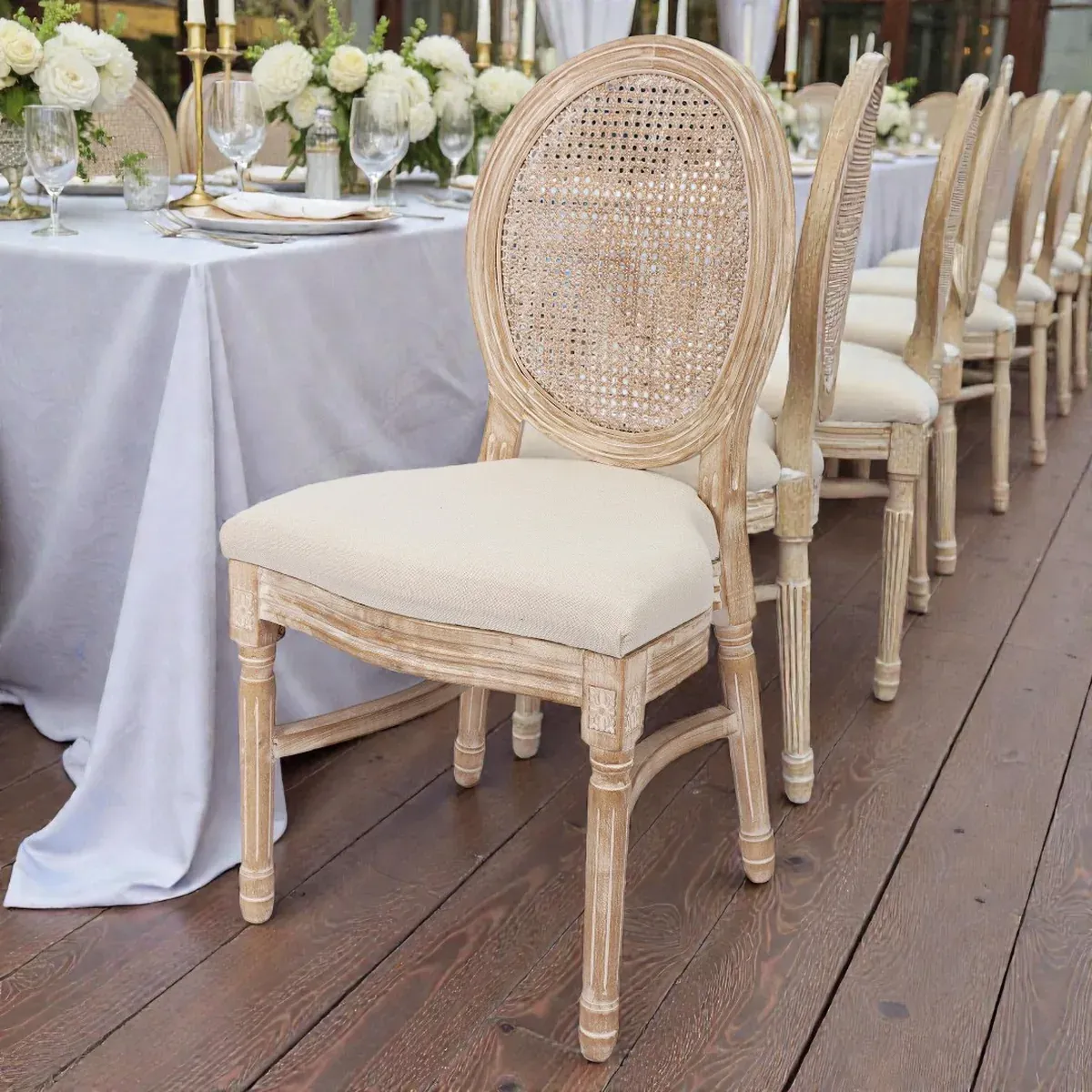 Reihe von Louis-Stühlen mit ovaler Rattan-Rückenlehne und cremefarbenem Polster an einer festlich gedeckten Tafel mit weißen Rosen